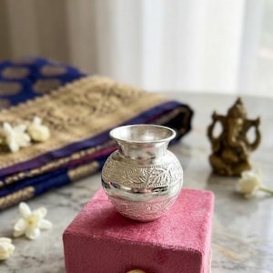 Könnte beinhalten: Eine kleine, silberfarbene Vase mit geprägtem floralem Design, die auf einer rosa Samtbox mit goldenem Verschluss steht. Die Vase hat eine breite Öffnung und einen abgerundeten Körper. Der Hintergrund umfasst gemusterten Stoff und eine kleine goldene Statue.