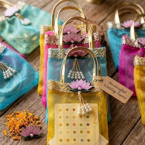May include: Colorful handcrafted wedding favor bags with gold handles, adorned with lotus flower embellishments and pearl accents. The bags are in shades of yellow, blue, and pink, with a tag that reads "Handcrafted Wedding Favors."
