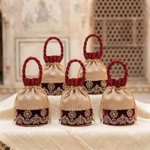 May include: Five drawstring pouches made of beige fabric with a dark red velvet base. Each pouch features a braided dark red handle and gold-toned embroidery in a floral pattern along the velvet section, finished with a gold trim border.