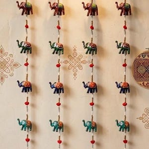 May include: Decorative wall hanging featuring rows of colorful elephant figurines. Each elephant is painted in shades of brown, green, blue, and teal, with intricate patterns. The elephants are strung together with beads and red accents, and hang from a wooden branch.