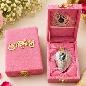 Puede incluir: Una caja rectangular de textura aterciopelada rosa con un cierre de metal dorado y el texto en hindi "Shubh Deepavali" en la tapa. En el interior, se muestra un colgante plateado con una piedra verde oscuro en forma de lágrima y cristales.
