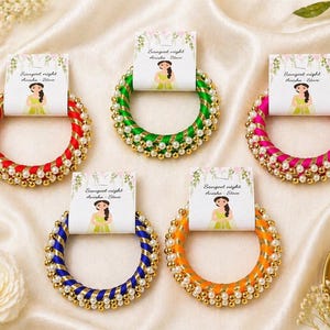 Op de afbeelding: Vijf kleurrijke armbanden met decoratieve parel- en gouden kraalaccenten. Elke armband heeft een kaart met een illustratie en de tekst "Sangeet night Anisha & Steve". De armbanden zijn rood, groen, roze, blauw en oranje.