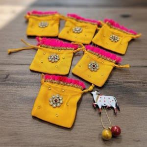 May include: Five small yellow drawstring pouches with bright pink fringe trim at the top. Each pouch features a central floral-shaped embellishment with clear stones and a red center. A white cow-shaped charm with red spots hangs from a gold cord.