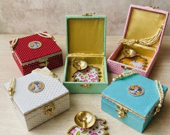 May include: Five square boxes in red, white, mint green, pink, and light blue, each featuring a pearl-beaded handle, a gold metal latch, and a central circular floral emblem. Inside the open boxes are gold metal diya lamps on floral-patterned bases.
