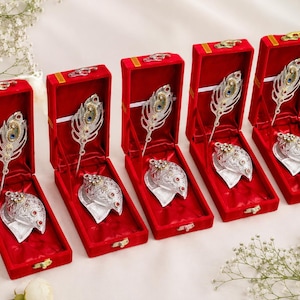 Puede incluir: Cinco cajas de regalo rectangulares de terciopelo rojo, cada una con un broche plateado en forma de pluma de pavo real y un adorno plateado a juego en forma de corazón. Los broches y adornos están decorados con pequeñas piedras decorativas azules, rojas y transparentes.