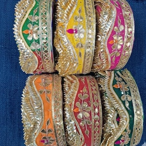 Op de afbeelding: Close-up van zes kleurrijke decoratieve stoffen linten. Elk lint heeft een centrale band in een andere kleur, waaronder groen, geel, roze, oranje, rood en groen. De linten zijn versierd met gouden metalen randen en bloemenborduursels.