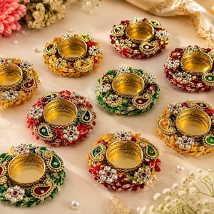 May include: A collection of ornate, colorful candle holders. Each holder features a gold-colored metal cup surrounded by a decorative ring of red, green, and gold accents, with white pearl-like embellishments. The candle holders are arranged on a light-colored surface.