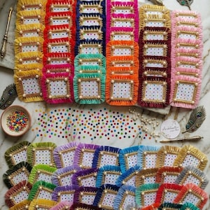 May include: A collection of colorful, square-shaped decorative items with a white dotted pattern, gold trim, and pearl accents. The items are arranged in rows and are surrounded by decorative elements, including peacock feathers and a small bowl of colorful beads.