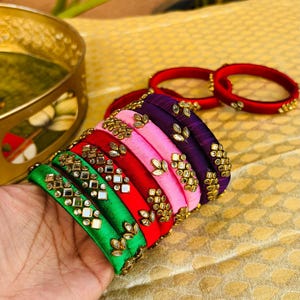 Puede incluir: Una colección de pulseras coloridas en tonos verdes, rojos, rosas y morados. Cada pulsera está adornada con adornos dorados. Las pulseras se exhiben sobre una superficie dorada texturizada, mostrando una vibrante exhibición de accesorios.