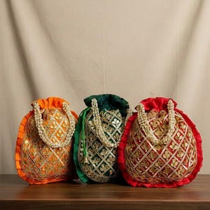May include: Three ornate drawstring bags in orange, green, and red, each with gold embroidery and a pearl handle. The bags are arranged on a dark wooden surface, showcasing their intricate designs and vibrant colors.