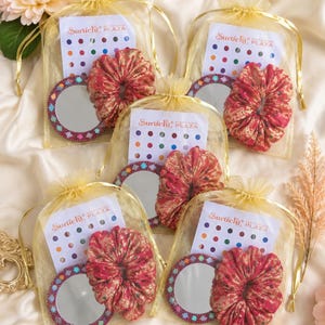 Peut inclure: Quatre sacs à cordon en organza doré contenant chacun un chouchou en tissu rouge et doré, un petit miroir rond à bordure géométrique colorée et une carte étiquetée Suruchi Plaza avec une grille de petits autocollants bindi multicolores.