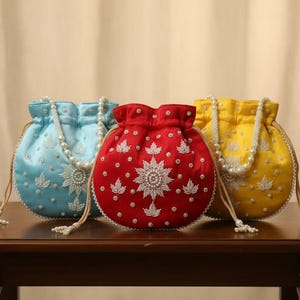 May include: Three handmade potli bags in light blue, red, and yellow, each adorned with white floral embroidery and a pearl beaded strap. The bags are displayed on a dark brown wooden table.