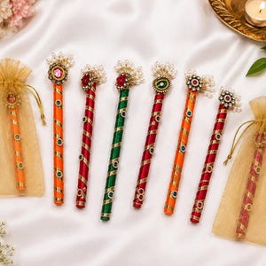 Könnte beinhalten: Eine Sammlung von sieben dekorativen Stäben, die mit leuchtend orangefarbenem, rotem und grünem Satinband mit Goldbesatz umwickelt sind. Jeder Stab hat oben eine Ansammlung weißer Kunstperlen und einen Edelstein, zwei Stäbe stecken in Organzabeuteln.