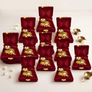 May include: A collection of ten open, deep red velvet jewelry boxes, each containing a gold-toned metal diya lamp with a leaf-shaped base and a small white floral accent. Two additional loose gold-toned metal diya lamps are placed separately.