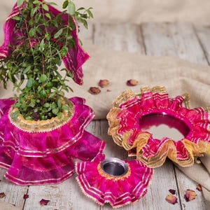 May include: A green plant in a pot covered with a bright pink ruffled fabric featuring gold trim, a circular mirror framed in matching pink and gold ruffled fabric, and a small metal container also adorned with the same pink and gold ruffled fabric.