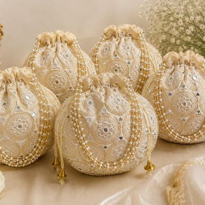 May include: Four round, cream-coloured drawstring pouches featuring intricate floral embroidery, small inset mirrors, and decorative pearl-beaded handles. Each bag is cinched at the top with matching fabric cords and finished with gold-toned metal tassels.