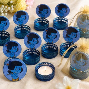 Könnte beinhalten: Eine Sammlung runder blauer Metallbehälter mit gitterartigen Seiten, die jeweils einen runden Deckel mit einer Cartoon-Illustration einer blauhäutigen Gottheit beim Flötenspiel haben. Ein Behälter enthält ein brennendes weißes Teelicht.