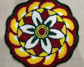 May include: A circular, floral-patterned rug with a vibrant design. The rug features a central flower with white petals, surrounded by red, yellow, and green accents. The outer edge is a dark green, creating a defined border. The rug is likely made of fabric.