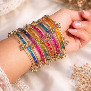 May include: A set of vibrant, multi-coloured bangles, featuring shades of blue, green, pink, and orange. The gold-coloured metal bangles are decorated with tiny, hanging bells. The jewellery is shown on a wrist, set against a soft, white backdrop.