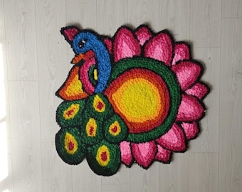 May include: A colorful, handmade rug featuring a peacock design. The peacock has a blue head and neck, a yellow beak, and a green and yellow tail. The rug is surrounded by a pink flower shape.