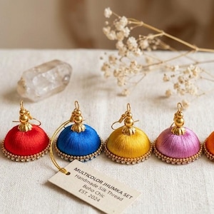 Peut inclure: Ensemble de six boucles d'oreilles en fil de soie faites à la main, aux couleurs vives : rouge, bleu, jaune, violet et orange. Chaque boucle d'oreille a un dessus doré et une base perlée. Une étiquette indique "Multicolor Jhumka Set, Boho Chic, Est. 2024."
