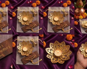 May include: A gold-toned metal lotus flower tea light candle holder with intricate cut-out petal designs. The set includes a small brass bell, a wooden plaque with the Hindi text "जय श्री राम" and "श्री हनुमान चालीसा", and a sheer drawstring pouch.