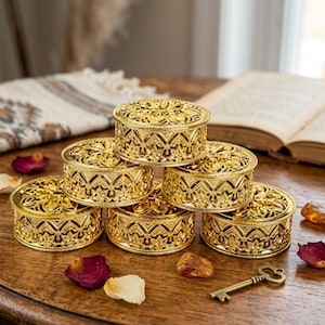 Può includere: Una collezione di sei scatole portagioie rotonde in metallo color oro con intricati motivi a filigrana intagliati sui coperchi e sui lati. Le scatole sono disposte in un gruppo, mostrando una finitura metallica e dettagliate lavorazioni.