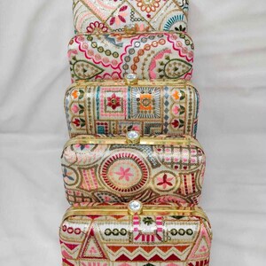 Op de afbeelding: Een stapel van vijf rechthoekige clutch-tassen van crèmekleurige stof met ingewikkeld, meerkleurig borduurwerk in tinten roze, oranje, groen en blauw. Elke tas heeft een goudkleurig metalen frame en een enkele doorzichtige kristallen sluiting.