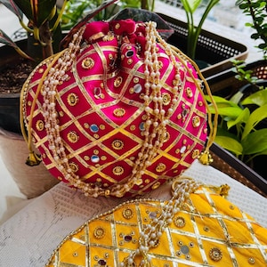 May include: A vibrant pink and gold embroidered pouch bag with a drawstring closure. The bag is adorned with gold trim, small mirrors, and pearl-like beads. A yellow bag with similar embellishments is also visible.