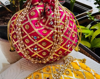 Könnte beinhalten: Eine leuchtend pinkfarbene und goldfarbene bestickte Beuteltasche mit Kordelzug. Die Tasche ist mit goldenen Verzierungen, kleinen Spiegeln und perlenartigen Perlen verziert. Eine gelbe Tasche mit ähnlichen Verzierungen ist ebenfalls sichtbar.