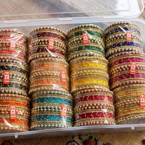 Könnte beinhalten: Eine transparente Plastikbox gefüllt mit Stapeln bunter Armreifen. Die Armreifen sind in verschiedenen Farben, darunter Rot, Orange, Grün und Blau, und mit kleinen, goldfarbenen Perlen verziert. Jeder Stapel ist in Klarsichtfolie verpackt.