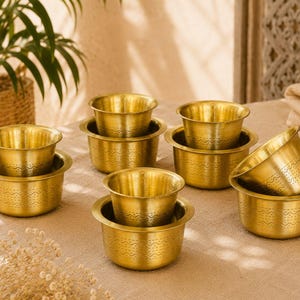 Può includere: Un set di quattro ciotole in ottone, ciascuna abbinata a una tazza in ottone più piccola inserita all'interno. Le ciotole e le tazze presentano una finitura dorata lucida e una fascia orizzontale con motivi geometrici in rilievo sui lati.