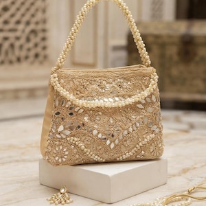 May include: A gold-toned handbag featuring intricate embroidery, small inset mirrors, and a handle crafted from rows of white pearls. The bag is adorned with detailed floral patterns and metallic thread work, complemented by a matching pearl-embellished accessory.