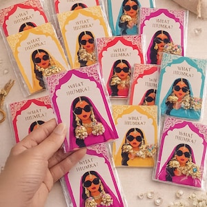 Può includere: Una collezione di cartoncini rettangolari con l'illustrazione grafica di una persona che indossa occhiali da sole e grandi orecchini jhumka. Ogni cartoncino riporta la scritta WHAT JHUMKA? e contiene un paio di orecchini jhumka color oro.
