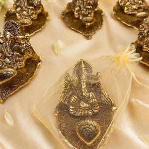 Op de afbeelding: Een verzameling metallic goudkleurige Ganesha-beeldjes, elk zittend op een gestructureerde bladvormige basis met een geïntegreerde kleine olielamphouder. Eén beeldje zit gedeeltelijk in een transparant organza geschenkzakje met trekkoord.