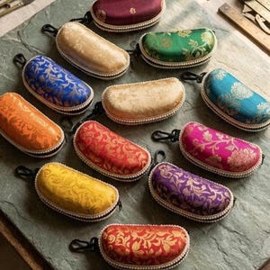 May include: A collection of colorful, patterned eyeglass cases. The cases are oval-shaped, with a zipper closure and a small loop. The cases are in various colors, including gold, blue, orange, red, yellow, purple, and pink, with floral designs.
