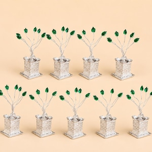 Può includere: Un set di nove alberi in miniatura in metallo color argento, ciascuno piantato in una base quadrata testurizzata color argento. Ogni albero presenta sottili steli di filo ramificato sormontati da piccoli accenti sfaccettati a forma di foglia verde brillante.
