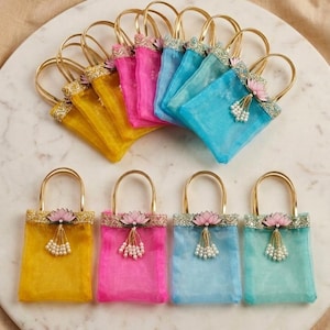 May include: A collection of small, colorful fabric bags with gold handles. Each bag is a different color, including yellow, pink, blue, and teal. They are decorated with a floral embellishment and dangling beads. The bags are arranged on a white marble surface.