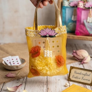 May include: A yellow mesh tote bag with gold handles, decorated with orange roses, a pink lotus flower, and a beaded tassel. The bag contains yellow flowers and a card with colored dots. A sign reads "Handcrafted Festive Favors."