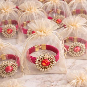 May include: A collection of red bracelets with gold clasps and ornate, jewel-toned centerpieces. Each bracelet is packaged in a sheer, cream-colored drawstring bag, creating a luxurious presentation. The bracelets are displayed on a marble surface.