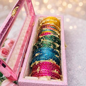 Puede incluir: Una colección de pulseras coloridas en una caja rosa con purpurina. Las pulseras son en tonos rosa, azul, verde y dorado. La caja tiene una tapa transparente y está forrada con una tela blanca suave. Una bonita idea de regalo.