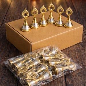 Peut inclure: Une collection de petites cloches en métal avec des sommets dorés et des bases argentées, chacune dotée d'une poignée décorative en forme de feuille. Six cloches sont posées sur une boîte en carton, d'autres sont dans un sachet plastique.