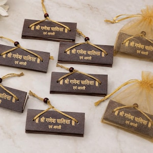 May include: A collection of rectangular dark brown booklets with gold text reading Shri Ganesh Chalisa and Aarti. Each booklet features a gold string with a small dark bead for hanging. Two booklets are enclosed in sheer gold organza drawstring bags.