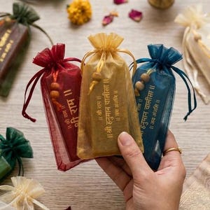 Puede incluir: Tres pequeñas bolsas transparentes con cordón en rojo, dorado y azul, cada una con texto dorado y un adorno de cuentas. Las bolsas están sobre una superficie de madera clara, con flores y una vela encendida en el fondo.