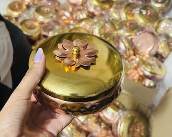 May include: A collection of gold and copper-toned decorative containers. One container is held in a hand, showcasing a flower-shaped embellishment on the lid. The containers have a hammered texture and are likely used for storage or display.