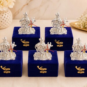 May include: Five silver-coloured metal figurines depicting a deity standing beside a cow, each mounted on a square base. These figurines are displayed on top of individual square, deep blue velvet gift boxes with gold-toned metal latches.