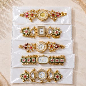 Puede incluir: Una vista cenital muestra cinco relojes dorados ornamentados, cada uno con diseños únicos. Los relojes presentan detalles intrincados con piedras preciosas rojas y transparentes, y varias formas de esfera. Se exhiben en embalajes blancos, sugiriendo un tema de joyería o accesorios.