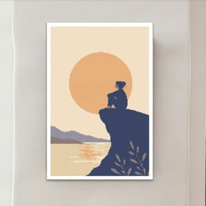 Op de afbeelding: Een ingelijste kunstdruk met een gepixeleerde illustratie van een persoon die op een klif zit en naar een grote oranje zon kijkt die ondergaat boven het water. Het kleurenpalet omvat tinten blauw, oranje en beige.
