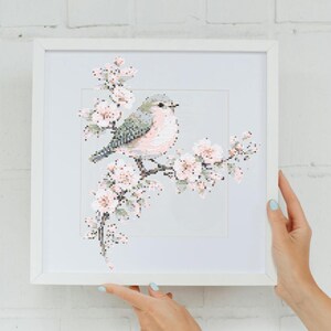 Op de afbeelding: Een ingelijst kruissteekkunstwerk met een vogel op een bloeiende tak. De vogel heeft een roze borst en grijze vleugels, en de bloemen zijn zachtroze. De witte lijst wordt door twee handen vastgehouden. Een delicaat kunstwerk.