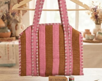 Puede incluir: Un bolso tote acolchado con rayas verticales que alternan tonos marrones y rosas. El bolso tiene dos asas de tela a juego y una abertura superior. Una etiqueta marrón rectangular con el texto HAADICREATIONJAIPUR se encuentra frente al bolso.